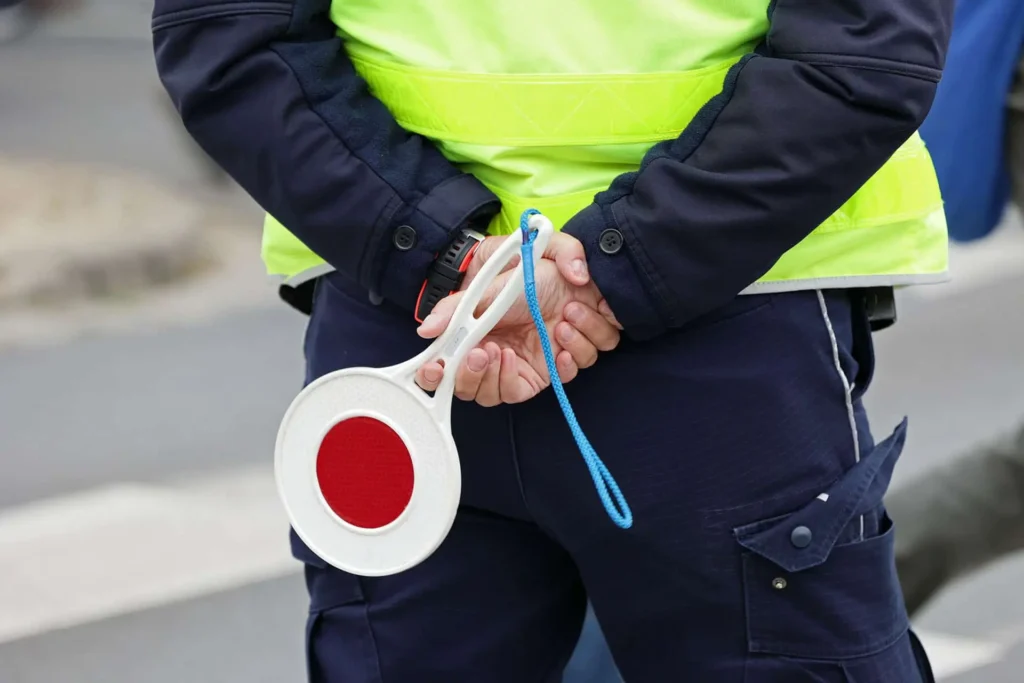 Ein Sicherheitsmitarbeiter in Uniform hält seine Haende hinter dem Ruecken verschraenkt, in einer Hand haelt er eine rote Signalpfeife.